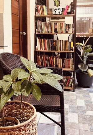 A wicker chair with a potted plant, a bookshelf filled with books, and a door in the background. - Little Garden Home | Garden View (Parañaque)