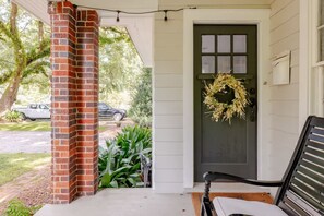 A porch with a brick pillar, a dark door with a wreath, and a bench. - Quaint little cottage nestled in the heart of East Hill! (Pensacola)