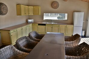 Cozinha compacta com micro-ondas, pia e janela acima do balcão. Cadeiras de vime e uma mesa estão em primeiro plano.
