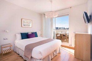A bedroom with a bed, a bedside table, a chair, a desk, a television, and a view of the city through a large window. - Elegant Eixample Penthouse with Private Terrace & City Views (Barcelona)