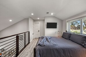 Quarto com cama, televisão presa à parede, janela com persianas e escada com corrimão.