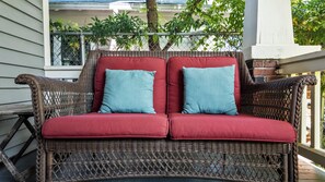 A wicker sofa with red cushions and two light blue pillows, positioned on a porch with a view of a house and greenery in the background. - Historic Borden Retreat: 2 Homes 6BR Hot Tub Sauna Pets OK (Galveston)