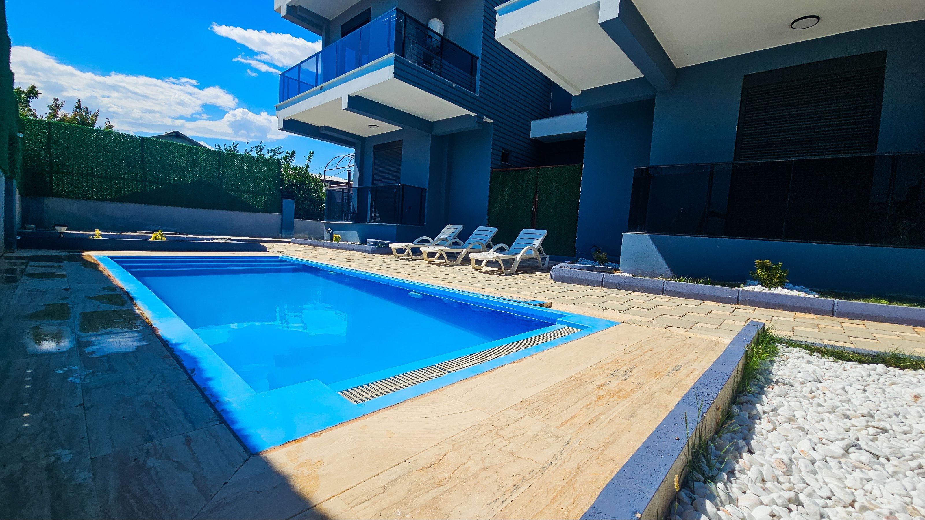 Una piscina moderna al aire libre con cubierta de madera, sillas de descanso y un edificio con balcones.