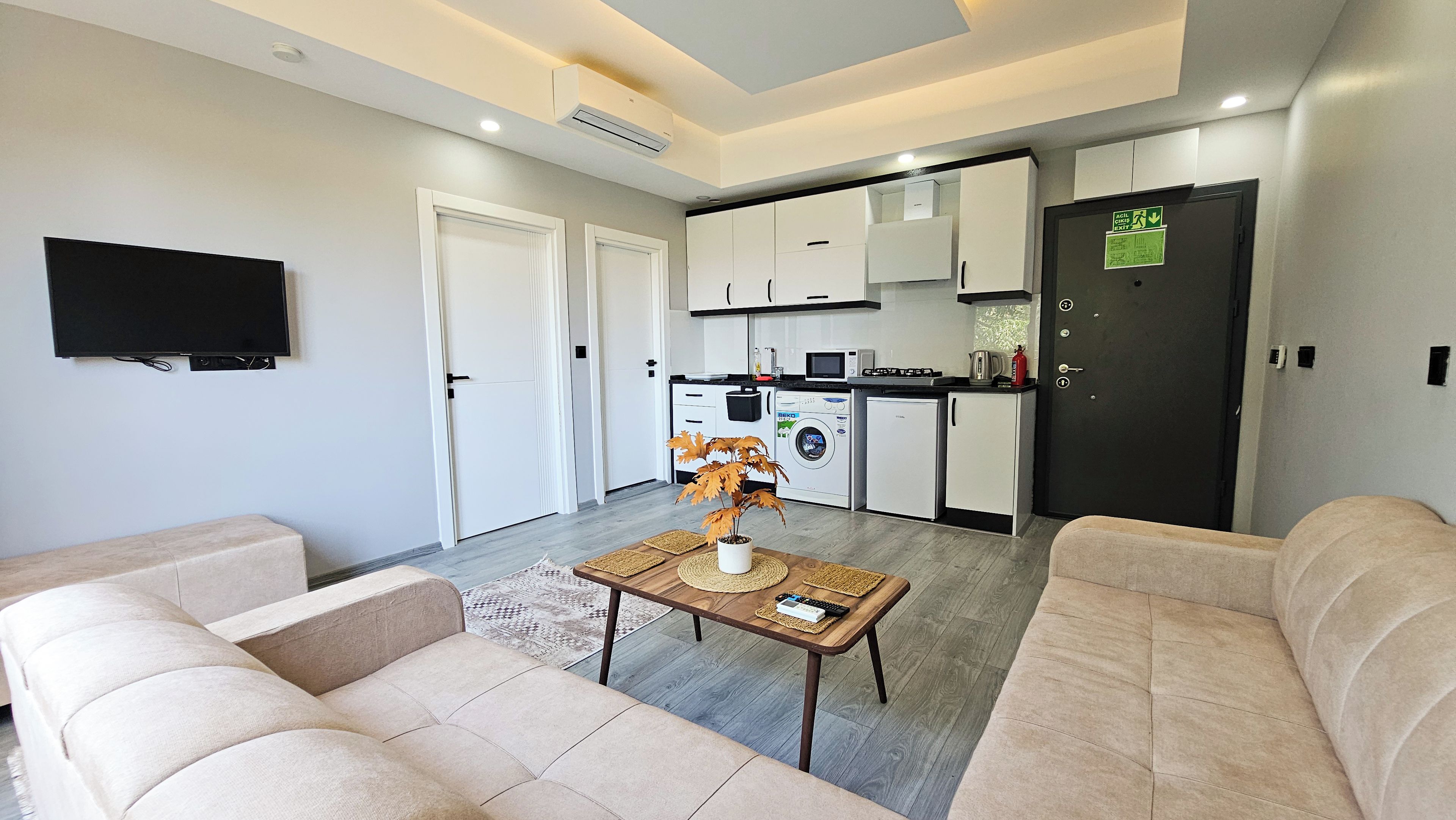 A modern living room with a flat-screen TV, beige sofas, a coffee table with decorative items, and a kitchen area with white cabinets, a microwave, and a refrigerator.