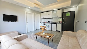 A modern living room with a flat-screen TV, beige sofas, a coffee table with decorative items, and a kitchen area with white cabinets, a microwave, and a refrigerator.