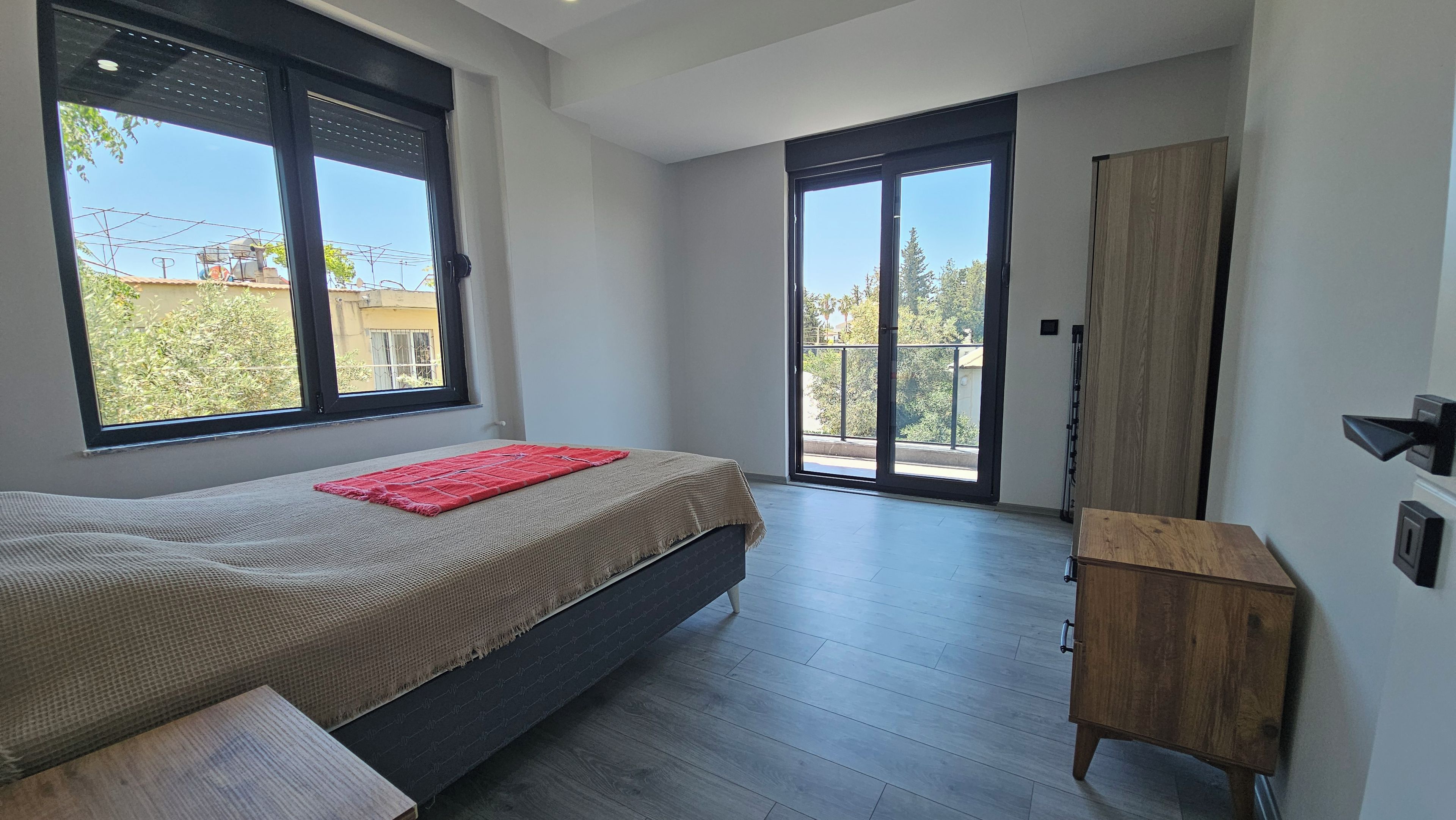 Un dormitorio con cama, un armario de madera y un ventanal con vistas a edificios y árboles.