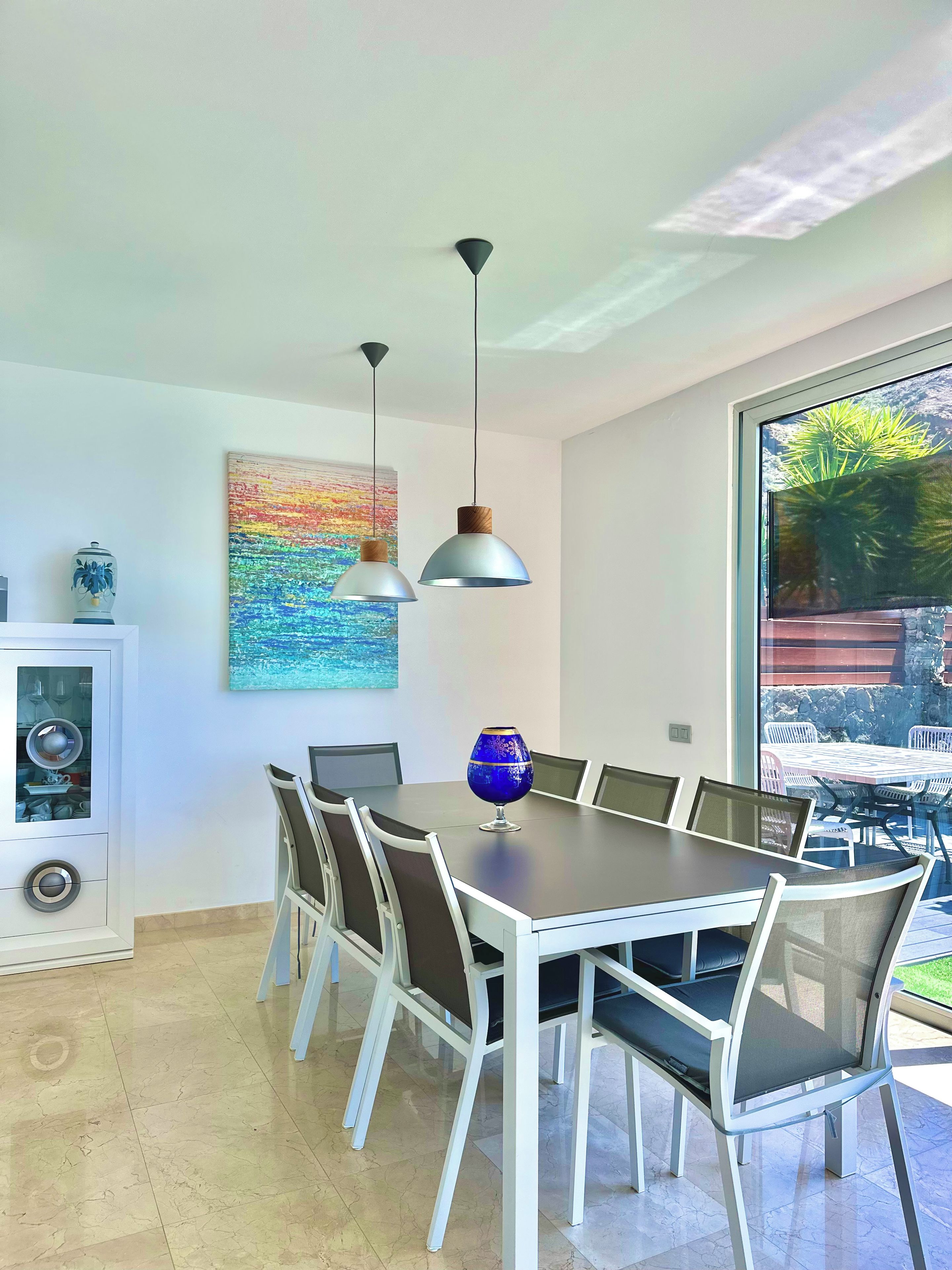 A dining area with a large table, chairs, pendant lights, a decorative vase, and a painting on the wall.