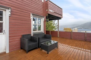 A wooden deck with wicker furniture and a glass table, overlooking a body of water and a distant mountain. - Tromsø 80 m² Apartment with balcony sea view (Tromsø)