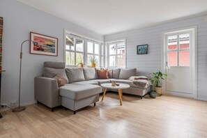 A modern living room with a grey sectional sofa, a round coffee table, and a large window with white shutters. - Tromsø 80 m² Apartment with balcony sea view (Tromsø)