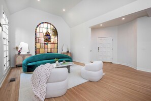 A spacious living room with a teal sofa, a white armchair, and a coffee table. A large stained glass window is visible in the background. - Alta Vista Terrace By Alora Reserve (Los Angeles)