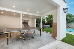 A ping pong table with a net, a small table, and a chair in an outdoor area with a pool and greenery. - Alta Vista Terrace By Alora Reserve (Los Angeles)