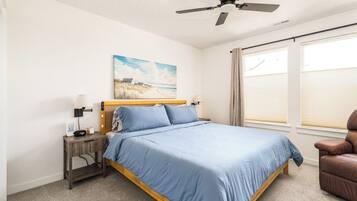 A bedroom with a bed, a nightstand, a lamp, a ceiling fan, a window with blinds, and a beach painting on the wall.