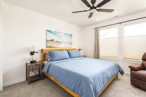 Une chambre à coucher avec un lit, une table de chevet, une lampe, un ventilateur de plafond, une fenêtre avec des stores et un tableau représentant une plage accroché au mur.