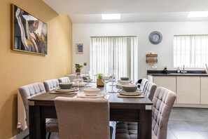 Spiseområde med et langt bord dækket til seks personer, omgivet af polstrede stole. Et køkken med vask og skabe ses i baggrunden.