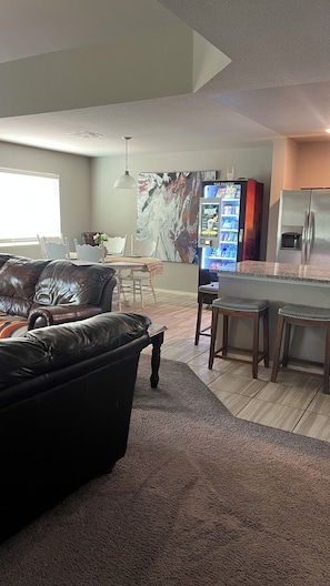 A living room with a black leather sofa, a dining area with a table and chairs, a kitchen with a refrigerator, and a TV mounted on the wall.