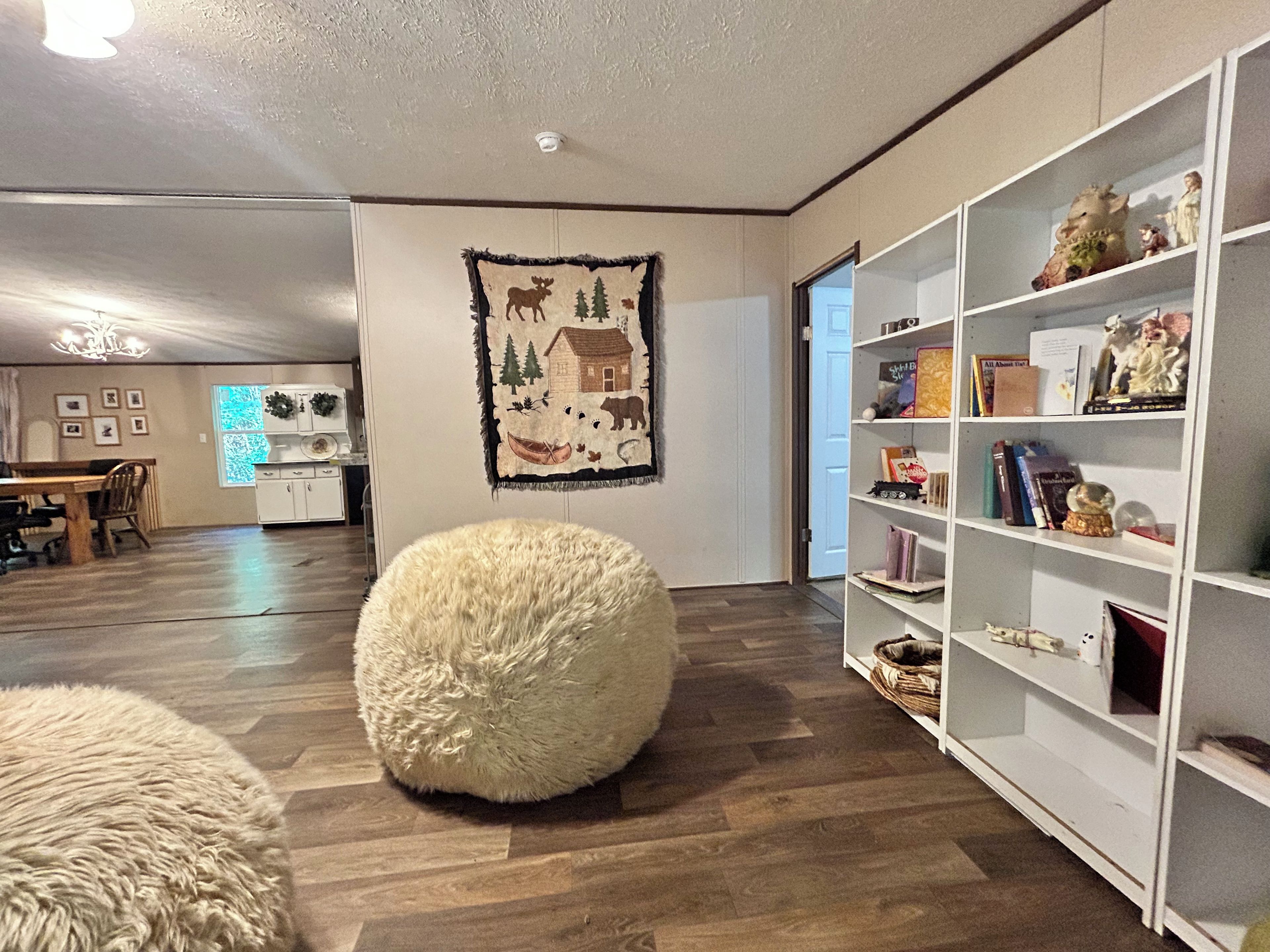 Een woonkamer met een houten vloer, een wit, zacht poefje, een boekenkast en een wand met een wandtapijt met natuurmotieven.