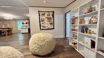 Een woonkamer met een houten vloer, een wit, zacht poefje, een boekenkast en een wand met een wandtapijt met natuurmotieven.