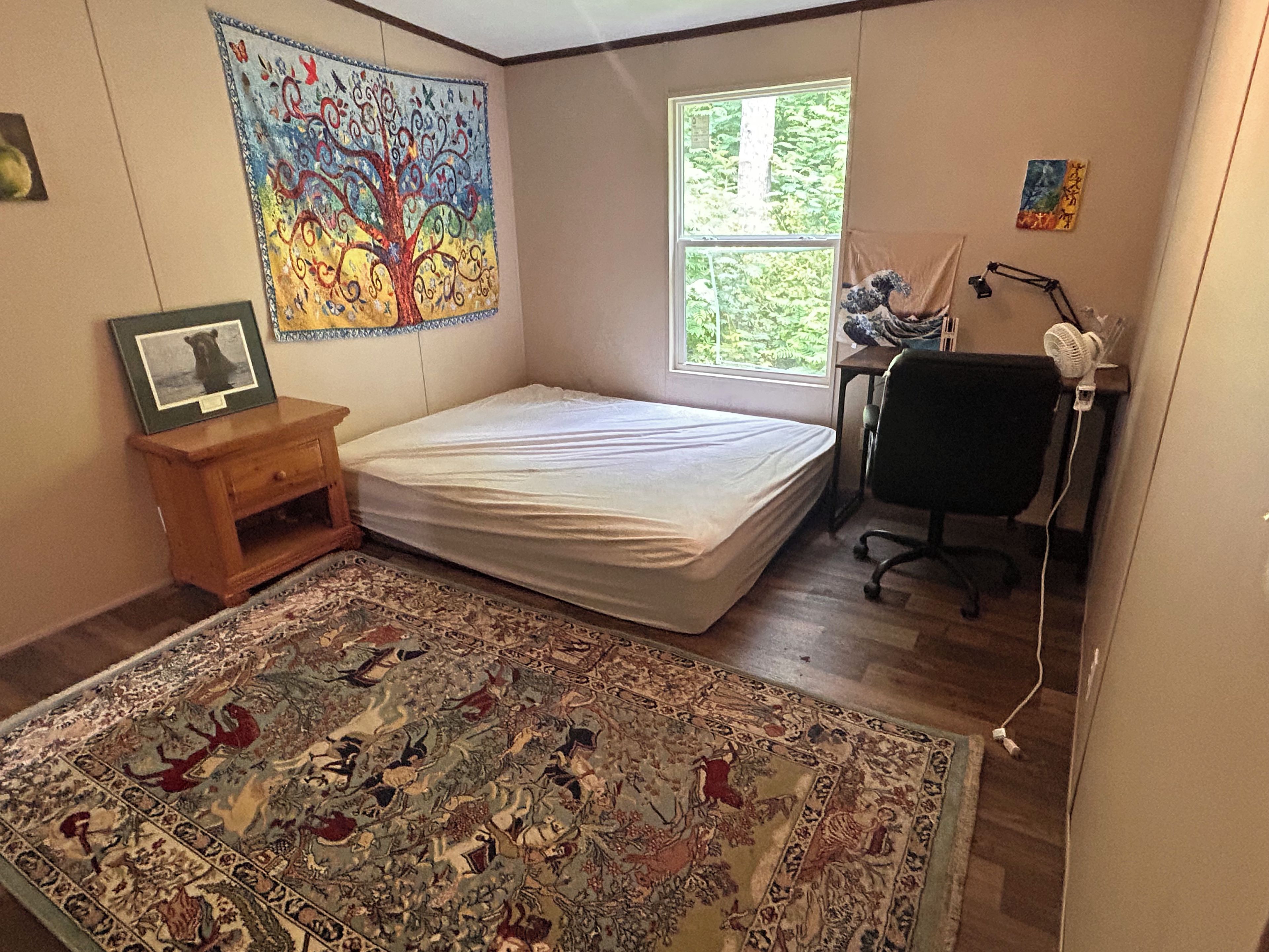 A bedroom with a bed, a desk, a chair, a window, a rug, and a wall painting.