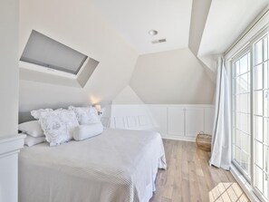 A bedroom with a sloped ceiling, a skylight, a bed with white linens, and a large window. - Harbor-View Condo w/ Sun Deck near Cape May (Cape May)
