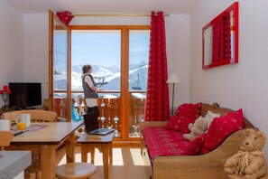 A room with a wooden sofa, a table with a laptop, a TV, and a view of mountains through a glass door. - Domaine skiable | Au pied du télésiège | Piscine (La Toussuire)