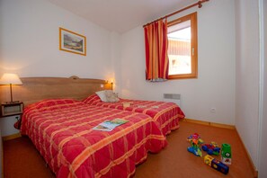 A hotel room with two beds, a nightstand, a lamp, a window with curtains, a radiator, and children's toys on the floor. - Domaine skiable | Au pied du télésiège | Piscine (La Toussuire)