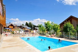 Un espace piscine avec des chaises longues et des parasols, entouré de bâtiments en bois et d’arbres.
