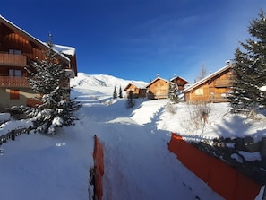 A snow-covered mountain village with wooden buildings and a clear blue sky. - Vue incroyable & exposition plein sud | Piscine (La Toussuire)
