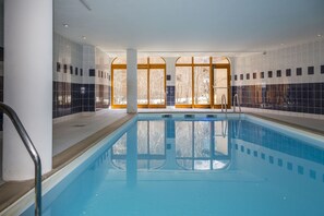 An indoor swimming pool with clear glass walls, a tiled floor, and a wooden door leading outside. - Au cœur du Parc de la Vanoise | Piscine sur place (Pralognan-la-Vanoise)