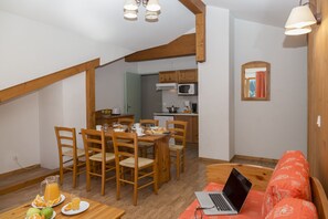 A dining area with wooden chairs and a table set for two, a kitchen with wooden cabinets, a microwave, and a window with curtains. - Au cœur du Parc de la Vanoise | Piscine sur place (Pralognan-la-Vanoise)
