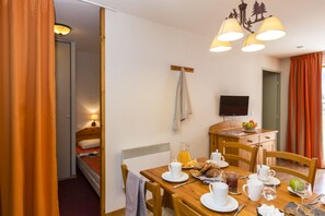 A hotel room with a dining table set for breakfast, a TV mounted on the wall, and a bed with an orange bedspread. - T2 à l’entrée de la station | Piscine couverte (Pralognan-la-Vanoise)
