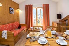 A cozy room with a wooden sofa, a dining table set with food and drinks, and a view of a mountain landscape through the window. - T2 à l’entrée de la station | Piscine couverte (Pralognan-la-Vanoise)