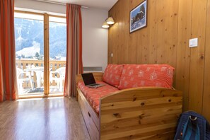 A wooden bed with a laptop, a window with curtains, a picture on the wall, and a view of mountains. - T2 à l’entrée de la station | Piscine couverte (Pralognan-la-Vanoise)