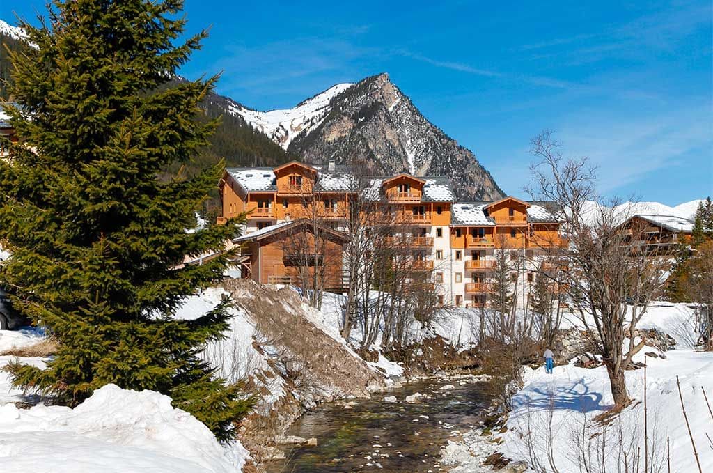Eine Berglodge mit mehreren Holzhäusern, einem Fluss und verschneiter Landschaft.