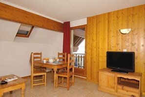 A room with a wooden dining table, chairs, a television on a wooden stand, and a balcony with a red curtain. - Télécabine Super Venosc à 2 pas | Station sportive (Les Deux Alpes)