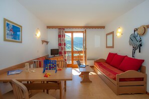 A cozy room with a wooden table, a red sofa, a television, and a view of the outdoors through a glass door. - Télécabine "Super Venosc" au pied de la résidence (Les Deux Alpes)