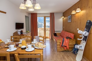 A dining area with a wooden table set for breakfast, a sofa with a snowboard, and a balcony with a view. - Piscine Intérieure | Au cœur du Parc de la Vanoise (Pralognan-la-Vanoise)
