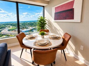 A dining area with a round table set for four, a potted plant, and a view of a cityscape through large windows. - Cityside Retreat | 2BR | Two Queen Beds (Birmingham)