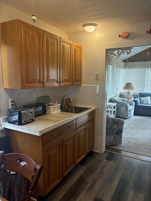 A compact kitchen with wooden cabinets, a countertop, and a dining area with a chair. - Fenced Beach House w/ Deck & BBQ | Nr Delaware Bay (Villas)