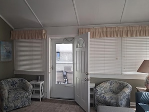Ein Zimmer mit zwei gemusterten Sesseln, einer zur Außenseite führenden Tür und einem Fenster mit Jalousien.