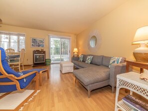 Ein Wohnzimmer mit einer Couch, einem Sessel und einem kleinen Tisch. Ein Fenster mit Jalousien, eine Lampe und ein dekorativer Spiegel sind an der Wand angebracht.