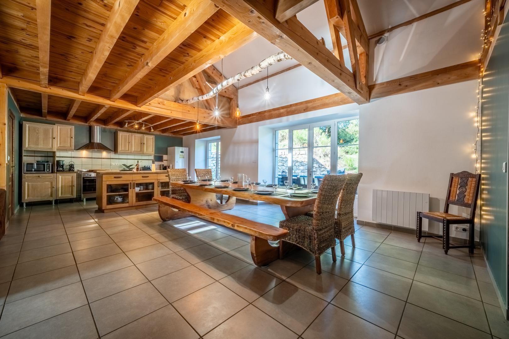 A spacious kitchen with wooden beams, a dining area with a large table and chairs, and a view of the outdoors through large windows.
