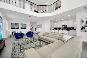 Ein geräumiges Wohnzimmer mit einem großen, beigefarbenen Ecksofa, einem Glastisch und blauen Stühlen. Man sieht eine Küche mit weißen Schränken sowie einen Essbereich mit einem weißen Tisch und Stühlen.