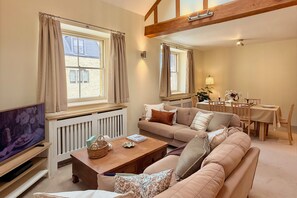 A cozy living room with a sofa, a coffee table, and a television. There are windows with curtains, a dining table with chairs, and a lamp. - Entire Home in Moreton-in-Marsh (Moreton-in-Marsh)
