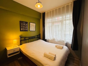 Ein Hotelzimmer mit einem Bett, einem Nachttisch, einer Lampe, zwei gerahmten Pflanzenbildern und einem Fenster mit durchscheinenden Vorhängen.