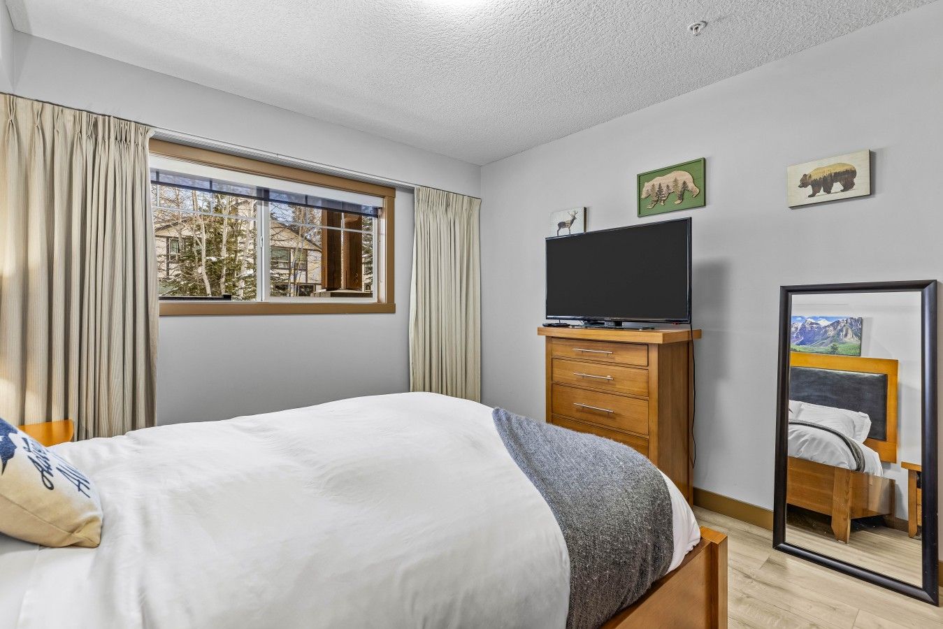 Une chambre à coucher avec un lit, un téléviseur posé sur un meuble en bois, une fenêtre avec des rideaux et un miroir qui reflète le lit.