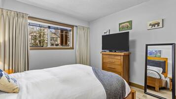 A bedroom with a bed, a television on a wooden stand, a window with curtains, and a mirror reflecting the bed.