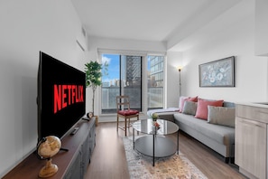 A modern living room with a flat-screen TV, a sofa, a coffee table, and a view of the city through large windows. - Heart of Downtown - 2BR - Near Dundas Square (Toronto)