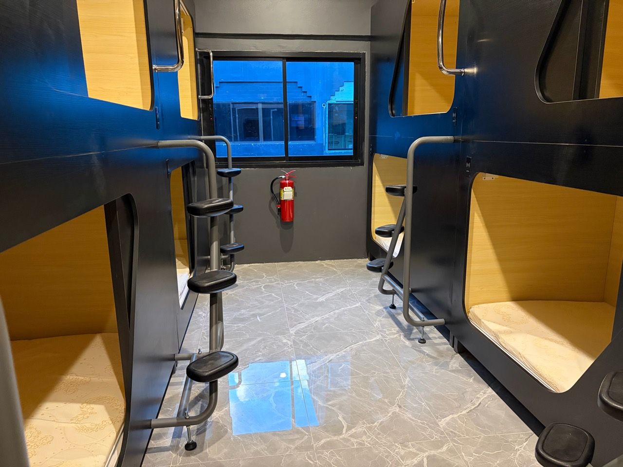 Un compartimento de tren con asientos de metal, un extintor y una ventana con vistas a edificios.