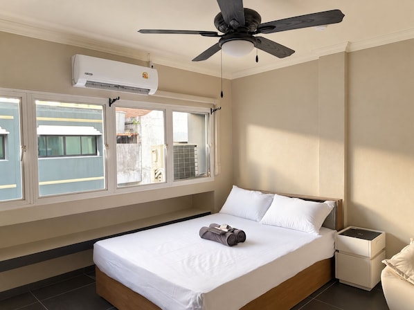 A hotel room with a bed, a ceiling fan, an air conditioning unit, a window with a view, and a bedside table with a lamp.