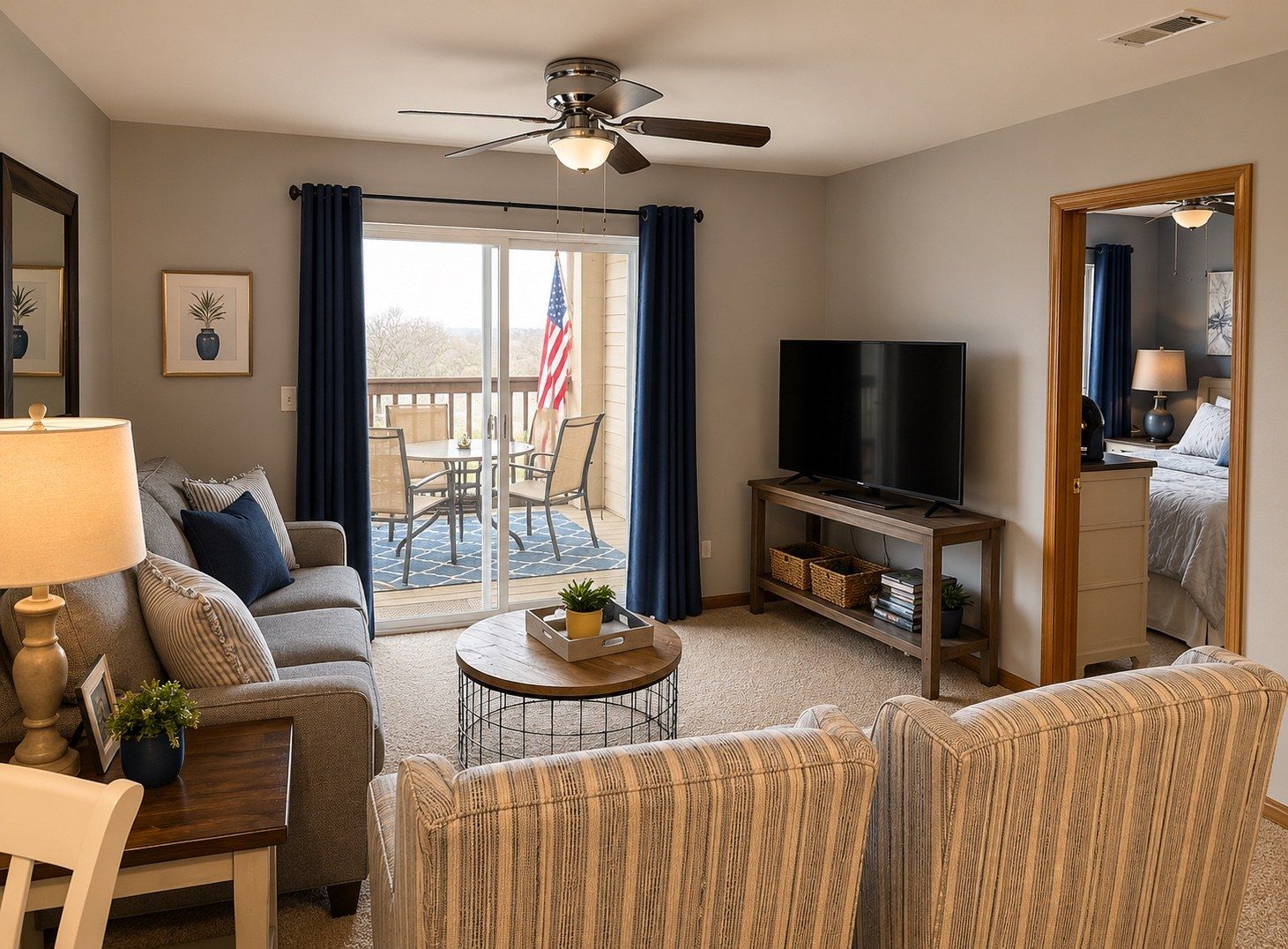 A living room with a sofa, armchair, coffee table, TV, and a balcony with an American flag.
